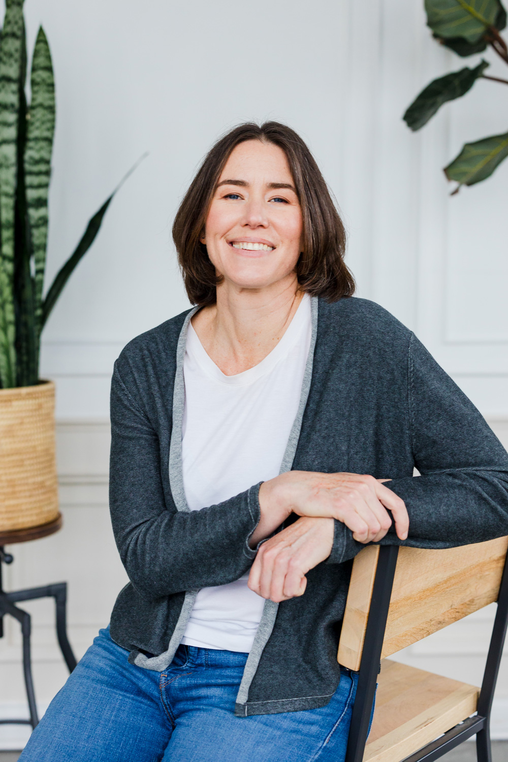 A woman with shoulder-length hair wearing a gray cardigan and white shirt sits on a wooden chair, smiling. She is in a bright room with a snake plant in a woven pot and another leafy plant in the background.