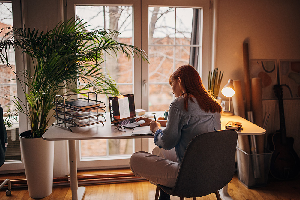 A person with long red hair sits at a desk in a home office, typing on a laptop. The room is warmly lit, with large windows showing trees outside. A potted plant, lamp, and stack of folders are on the desk. A guitar leans against the wall.