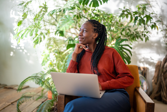 A person in a rust-colored shirt sits in a chair with a laptop on their lap, looking thoughtfully to the side. They are surrounded by lush green plants, creating a serene, contemplative atmosphere.