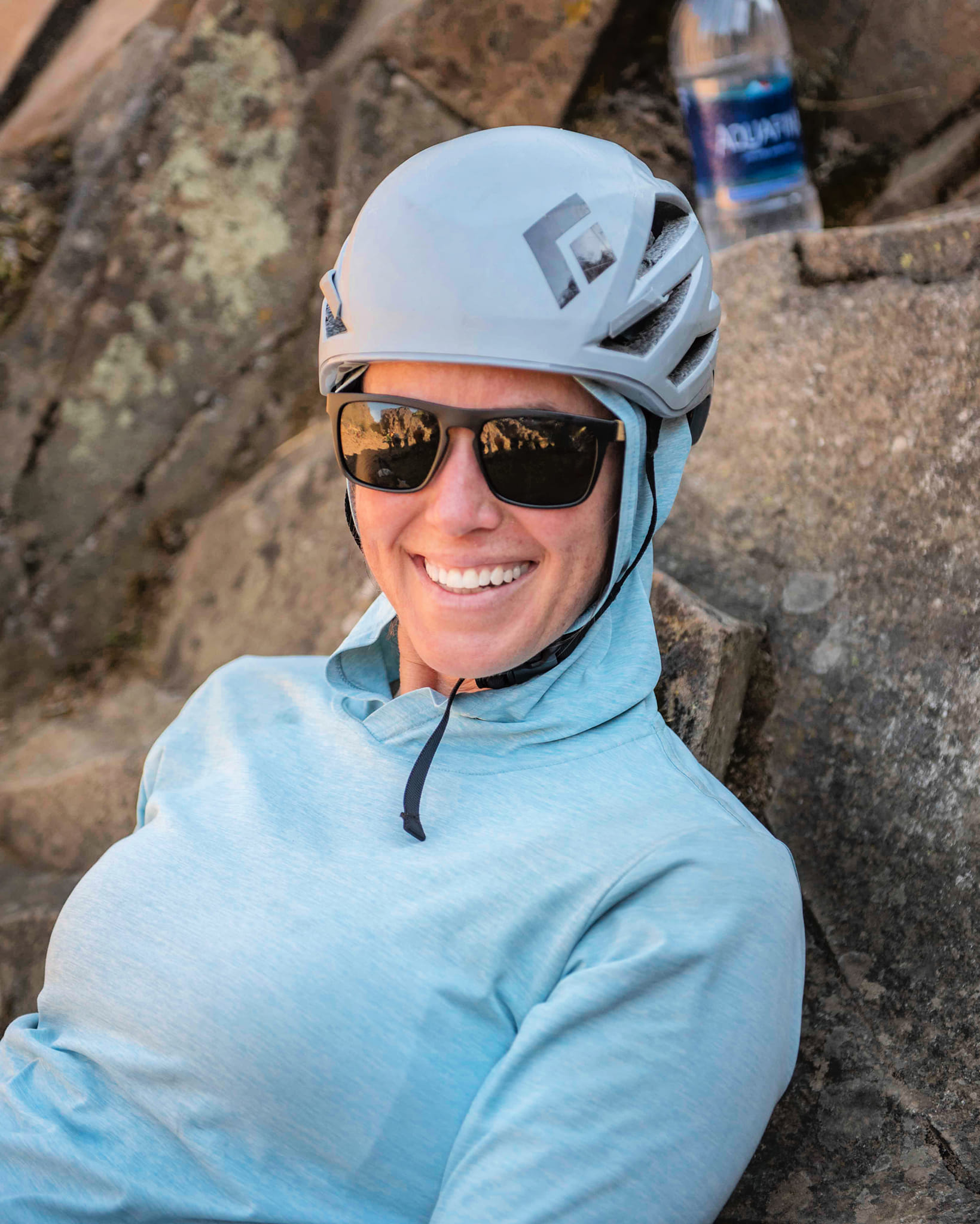 A person wearing a light blue hoodie, sunglasses, and a gray helmet smiles while sitting against a rock outdoors, with a water bottle visible in the background.